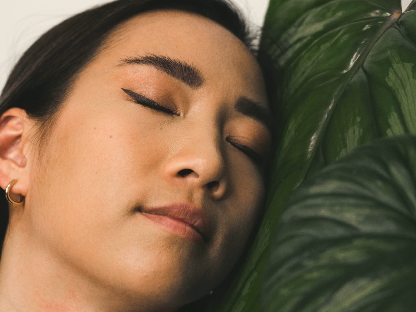 A face at rest, eyes closed, against a single deep green philodendron leaf