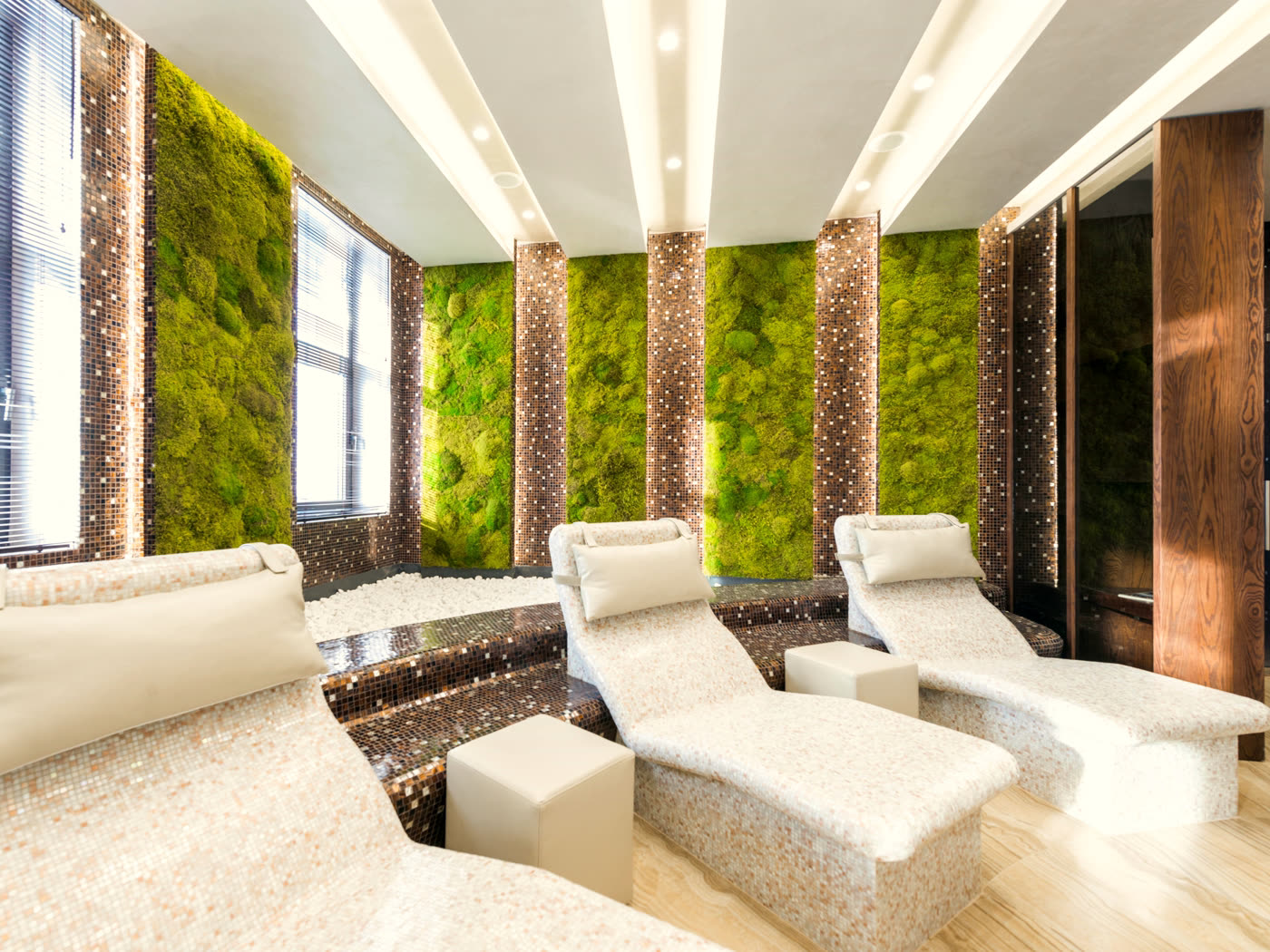 A quiet spa lounge: white stone loungers before a moss-green wall with bronze mosaic columns