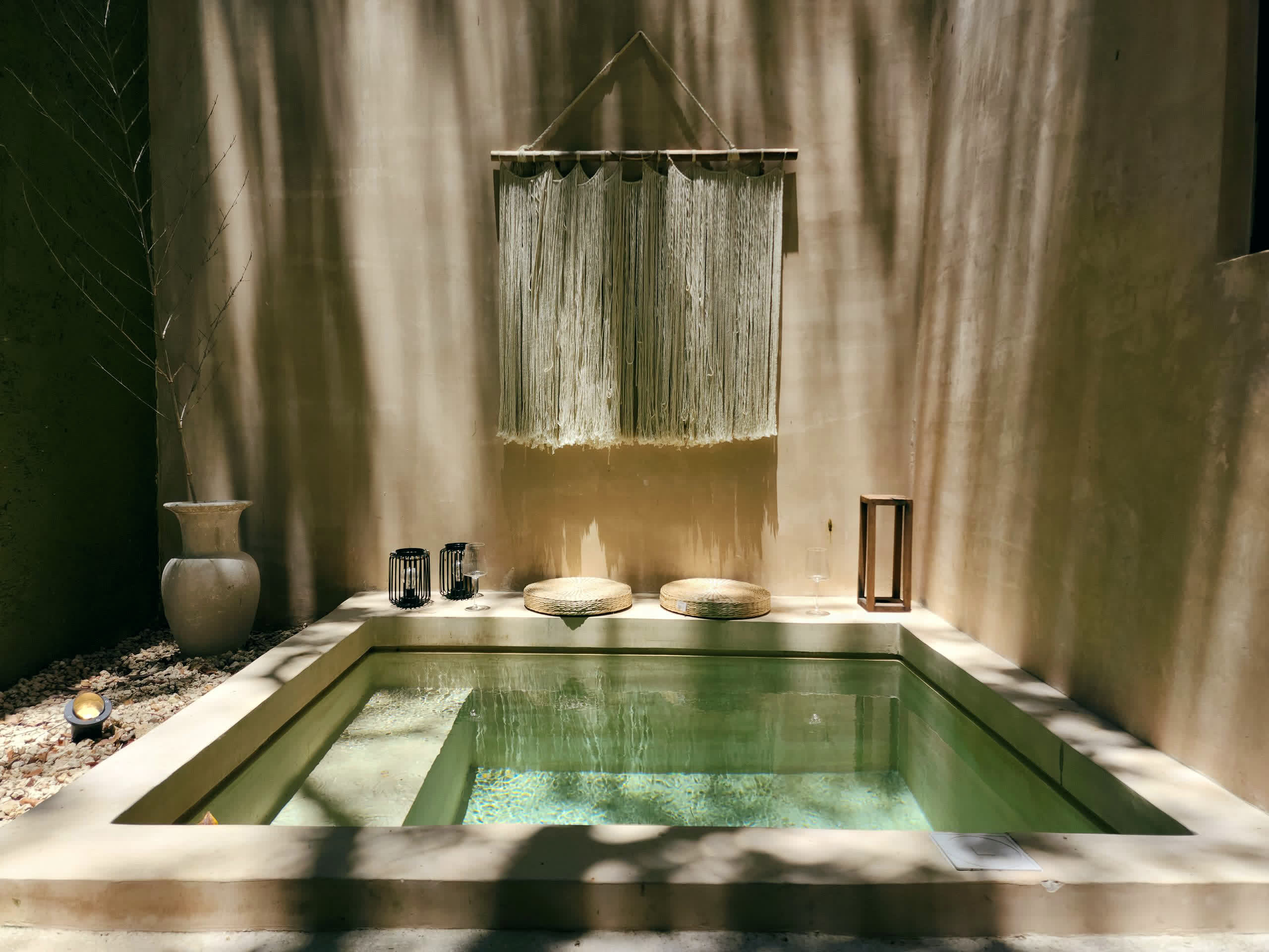 A sunlit plaster plunge pool with lanterns and a dried branch, empty and still.